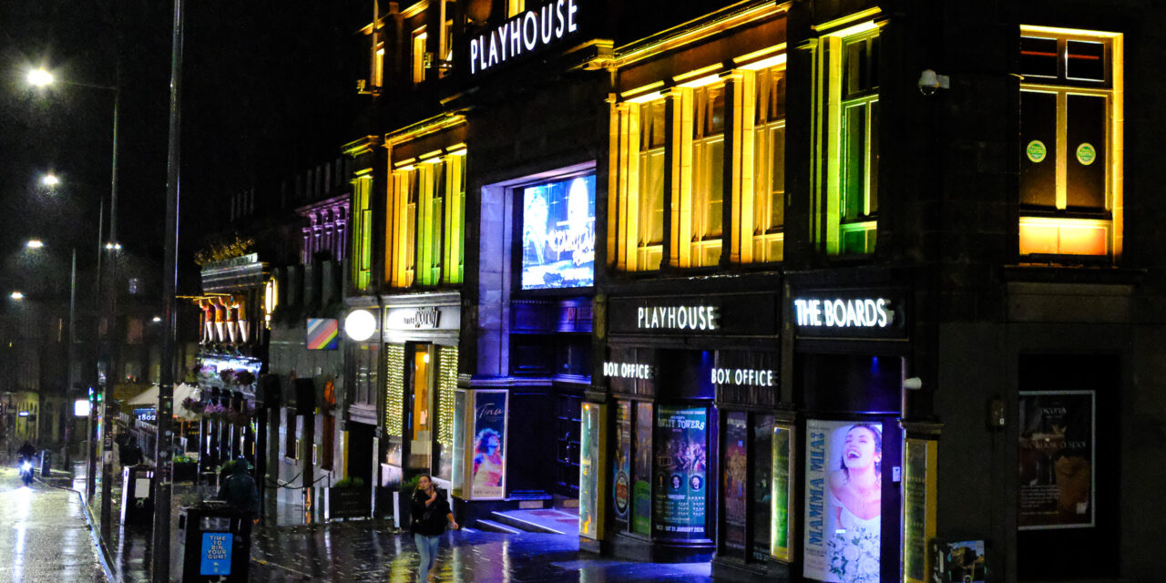 Night street scene with illuminated shops and wet pavement reflecting lights.